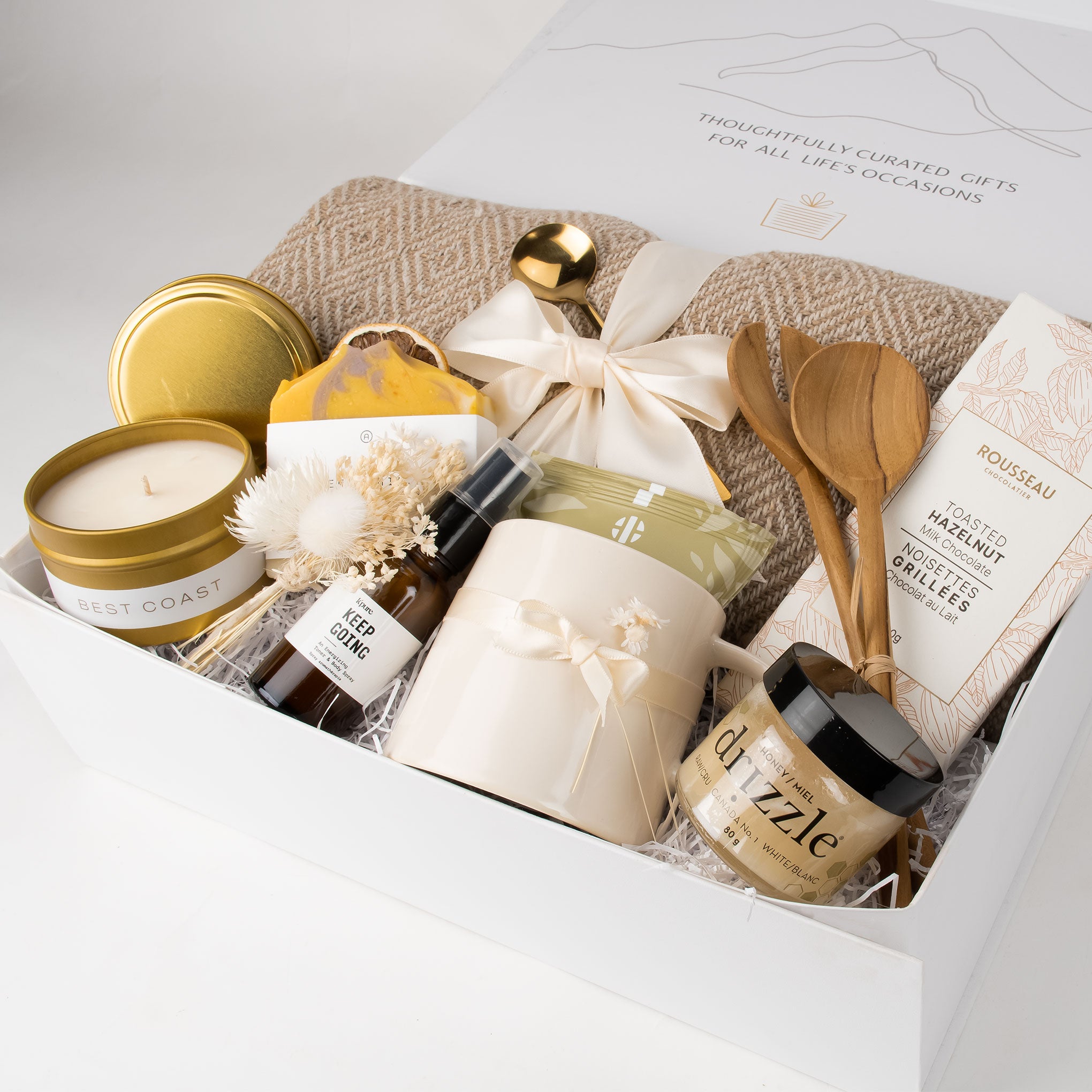 A curated gift box filled with premium items including a candle, mugs, wooden utensils, and packaged snacks, arranged in a white box with a ribbon.