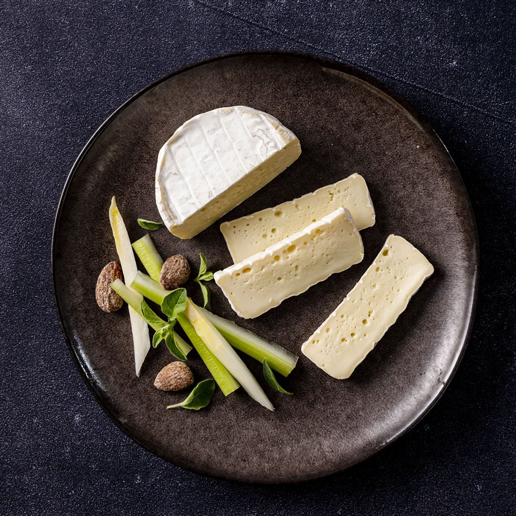 Four wedges of soft white cheese arranged on a dark round plate with sliced green vegetables and a few almonds on a dark textured background.