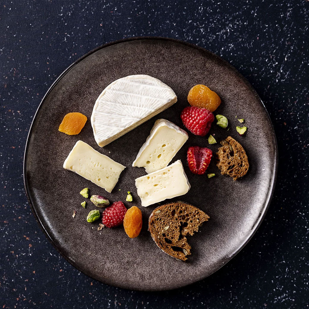 An overhead view of a dark round plate holding assorted cheeses, crackers, dried apricots, raspberries, and pistachios on a dark speckled background.