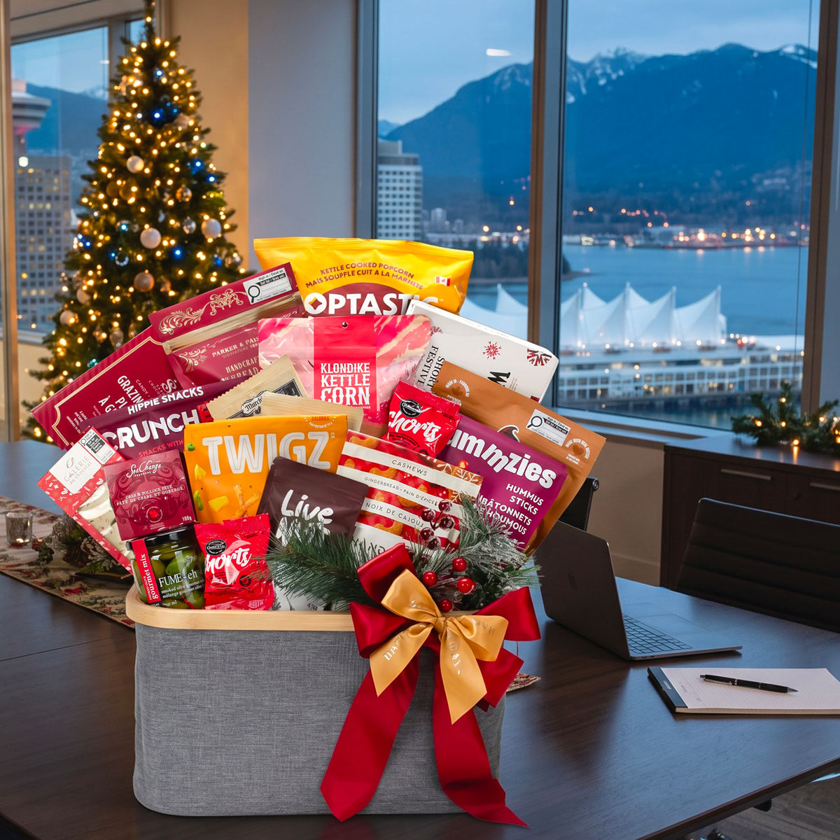 A gourmet Holiday gift basket filled with assorted snack bags and packaged food items, tied with a red and gold ribbon, placed on a desk in a modern office with large windows showing a city and Vancouver mountain view.