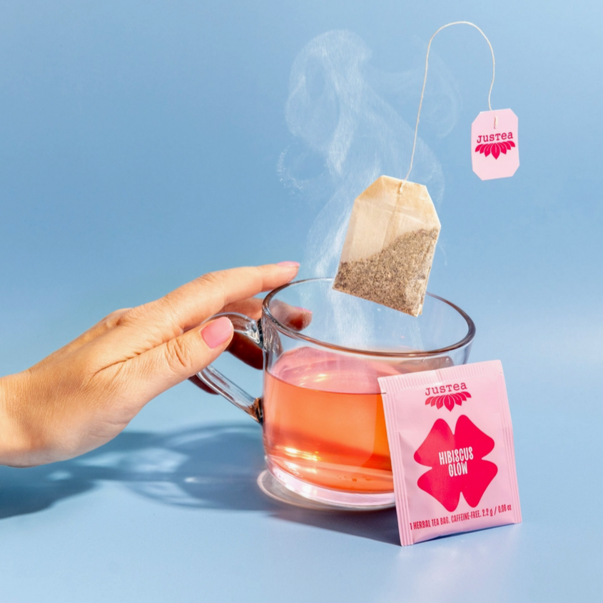 Hand holding a steaming glass of tea with a pink tea bag and box on a blue background