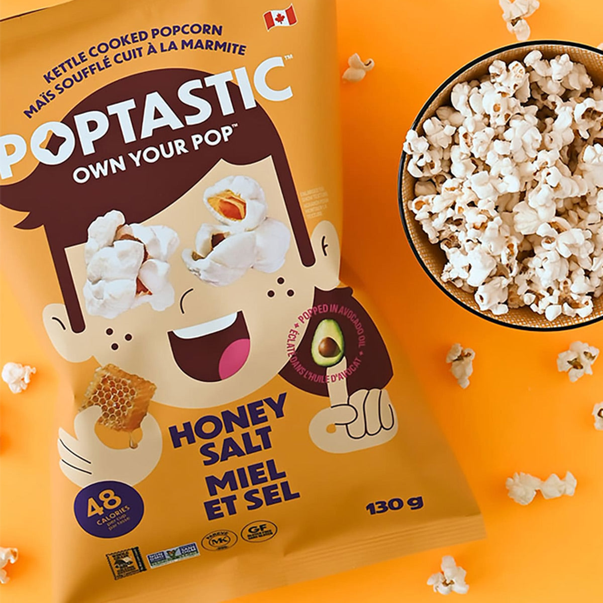 A bag of Poptastic Honey Salt kettle cooked popcorn sits on a bright orange background next to a bowl filled with popcorn pieces.