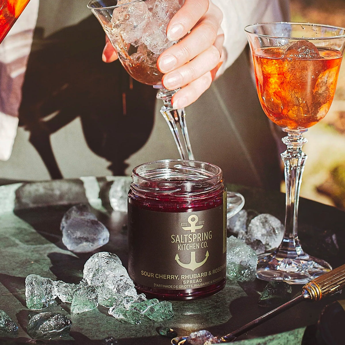 A jar of Saltspring Kitchen Co. Sour Cherry, Rhubarb & Rosehip Preserved Juice spread sits on a dark slate tray surrounded by ice cubes, with a hand pouring a dark red drink from a martini glass into it, and another filled glass standing nearby.