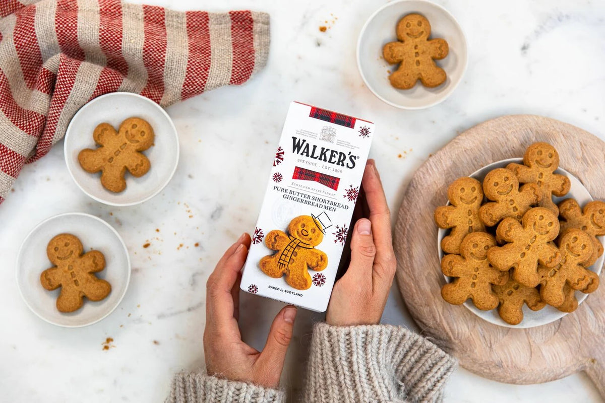 A person holds a box of Walker's Gingerbread Men in a cozy kitchen setting, surrounded by bowls of baked gingerbread cookies and a striped cloth on a white marble surface.