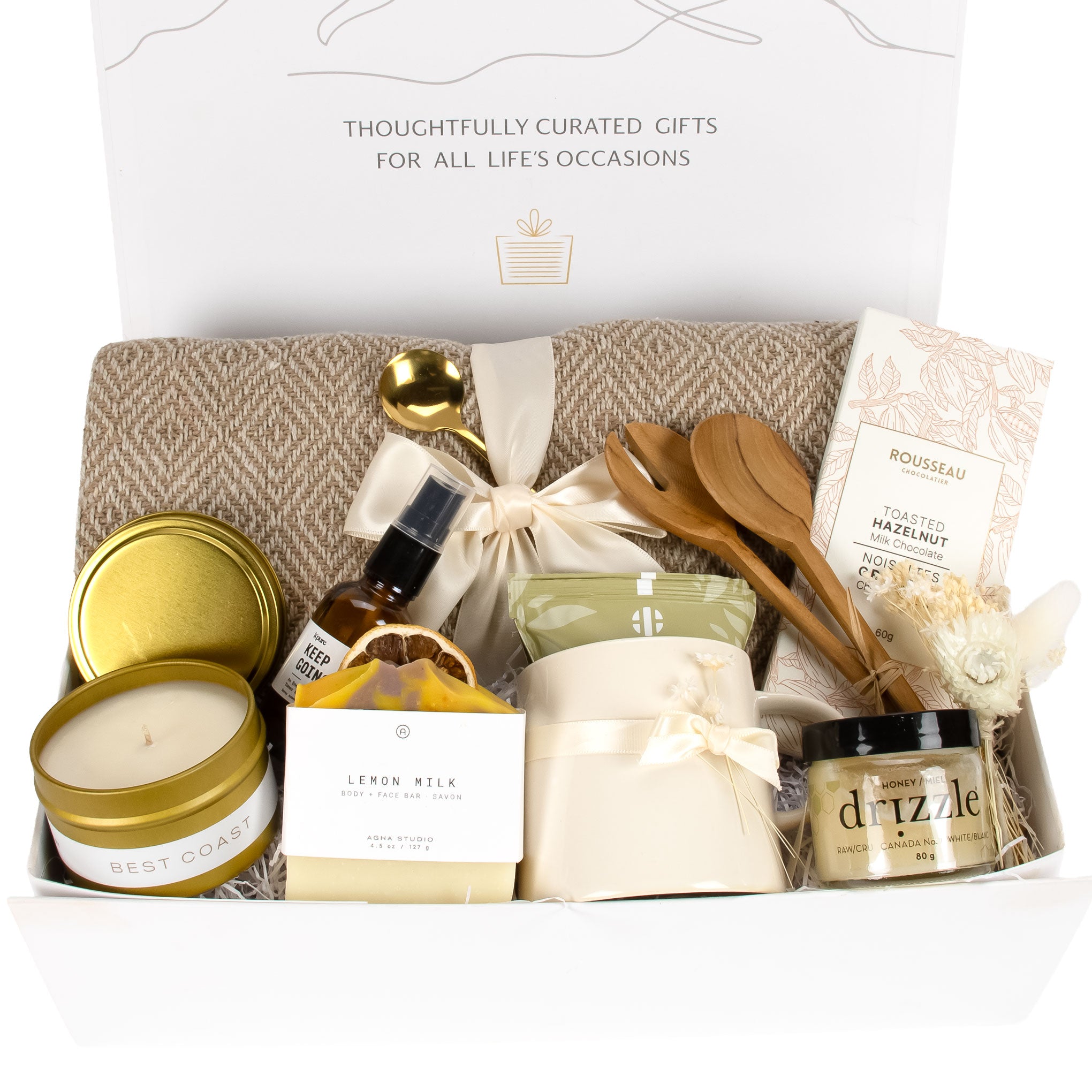 Open white Artisan Living gift box, beautifully arranged with a beige cotton throw, gold tin candle, Rousseau chocolate bar, ceramic mug with ribbon, and Drizzle honey jar.