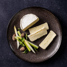Four wedges of soft white cheese arranged on a dark round plate with sliced green vegetables and a few almonds on a dark textured background.