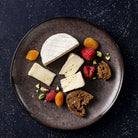 An overhead view of a dark round plate holding assorted cheeses, crackers, dried apricots, raspberries, and pistachios on a dark speckled background.