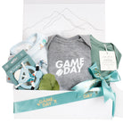 Game Day hockey baby gift box featuring a grey heathered bodysuit, sage green Pip and Phee bamboo onesie, Loulou Lollipop hockey bib, and a blue silicone mask teether in a white box with mountain line art.