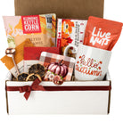 An autumn-themed gift box filled with seasonal items including a white ceramic mug with 'hello autumn' printed on it, a bag of Live Nuts, a box of Klondike Kettle Corn, dried citrus slices, a small pumpkin ornament, and fall-themed stationery inside a white and brown cardboard box.