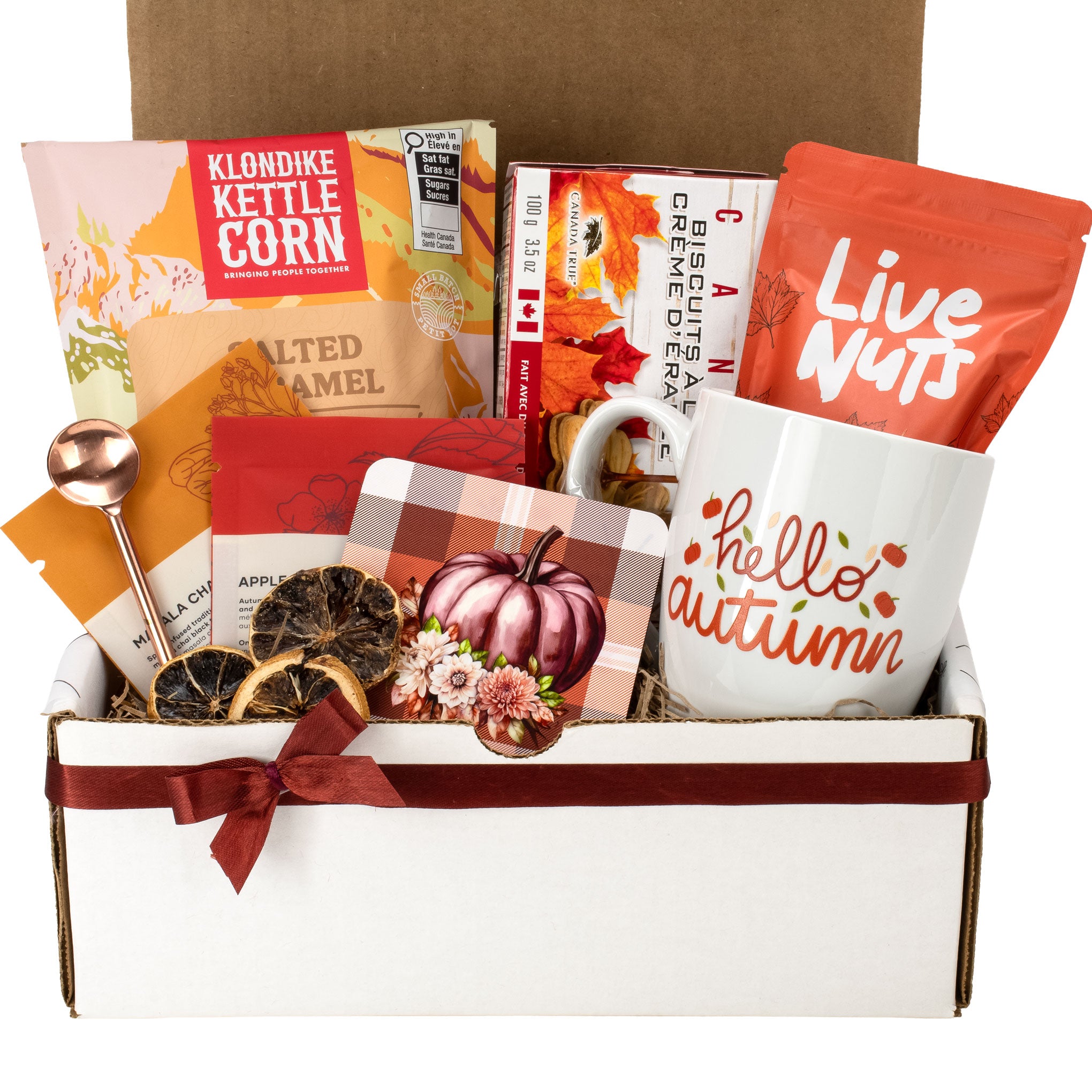 An autumn-themed gift box filled with seasonal items including a white ceramic mug with 'hello autumn' printed on it, a bag of Live Nuts, a box of Klondike Kettle Corn, dried citrus slices, a small pumpkin ornament, and fall-themed stationery inside a white and brown cardboard box.