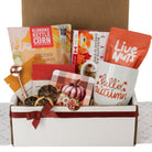 An autumn-themed gift box filled with curated treats and mugs. Visible items include a white ceramic mug printed with 'hello autumn' in orange, a bag of Live Nuts, a box of Klondike Kettle Corn, a bag of caramel-flavored snack, dried orange slices, a small rose-themed card, and a brown kraft gift box tied with a dark red ribbon.