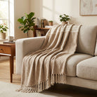 A beige textured throw blanket with fringe tassels draped over a light beige sofa in a warm, sunlit living room.
