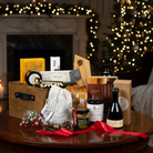 Gourmet gift basket with gourmet crackers, cheese, and other items on a table in front of a fireplace and Christmas tree.