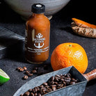 Bottle of Saltspring Kitchen Co. hot sauce on a dark surface with coffee beans and an orange.