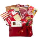 A red gift basket filled with assorted packaged snacks and treats, including Morris Salted Chocolate Bar, Parker & Priest Handmade Bread Cookies, Clonoke Kettle Corn, and a bag of mixed nuts, tied with a red ribbon on a white background.