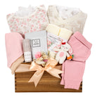 A baby gift basket arranged in a wooden crate with a peach ribbon bow, containing a floral baby bodysuit, a cream patterned top, pink swaddle blankets, a small plush bunny toy, folded pink clothing, and packaged baby care items on a white background.