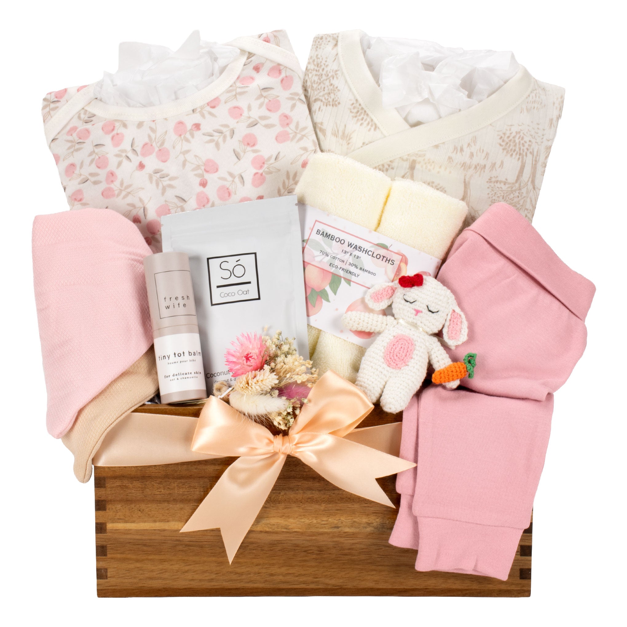 A baby gift basket arranged in a wooden crate with a peach ribbon bow, containing a floral baby bodysuit, a cream patterned top, pink swaddle blankets, a small plush bunny toy, folded pink clothing, and packaged baby care items on a white background.
