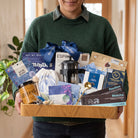 A person in a green sweater holds a wooden gift tray filled with assorted tea and coffee gift items, including a French press, blue ribbon, bags of Rooibos and Matcha, and packaged tea products, in an indoor setting.