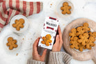 A person holds a box of Walker's Gingerbread Men in a cozy kitchen setting, surrounded by bowls of baked gingerbread cookies and a striped cloth on a white marble surface.