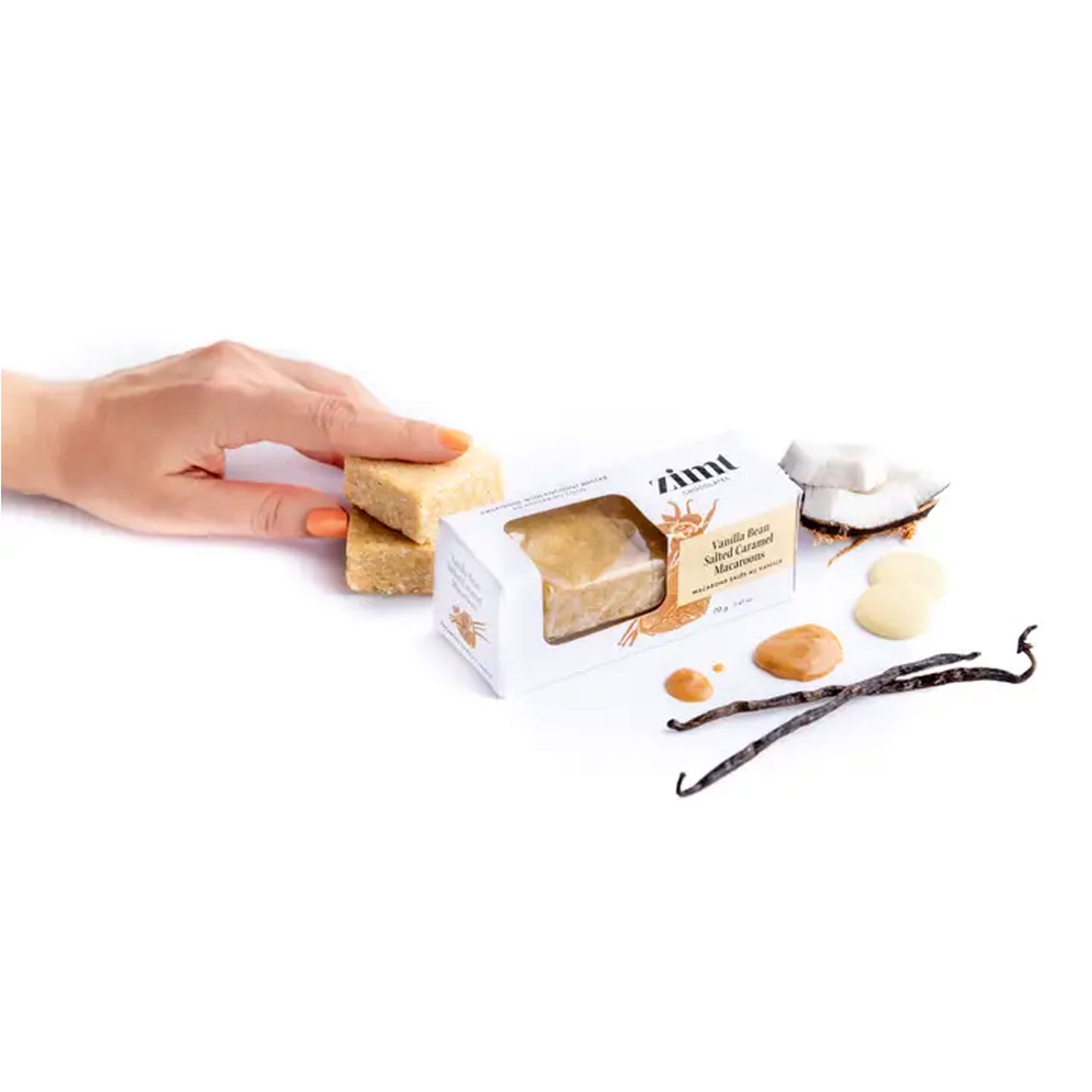 Hand holding a vegan macaroon next to a  Zimt box with a coconut and vanilla bean on a white background