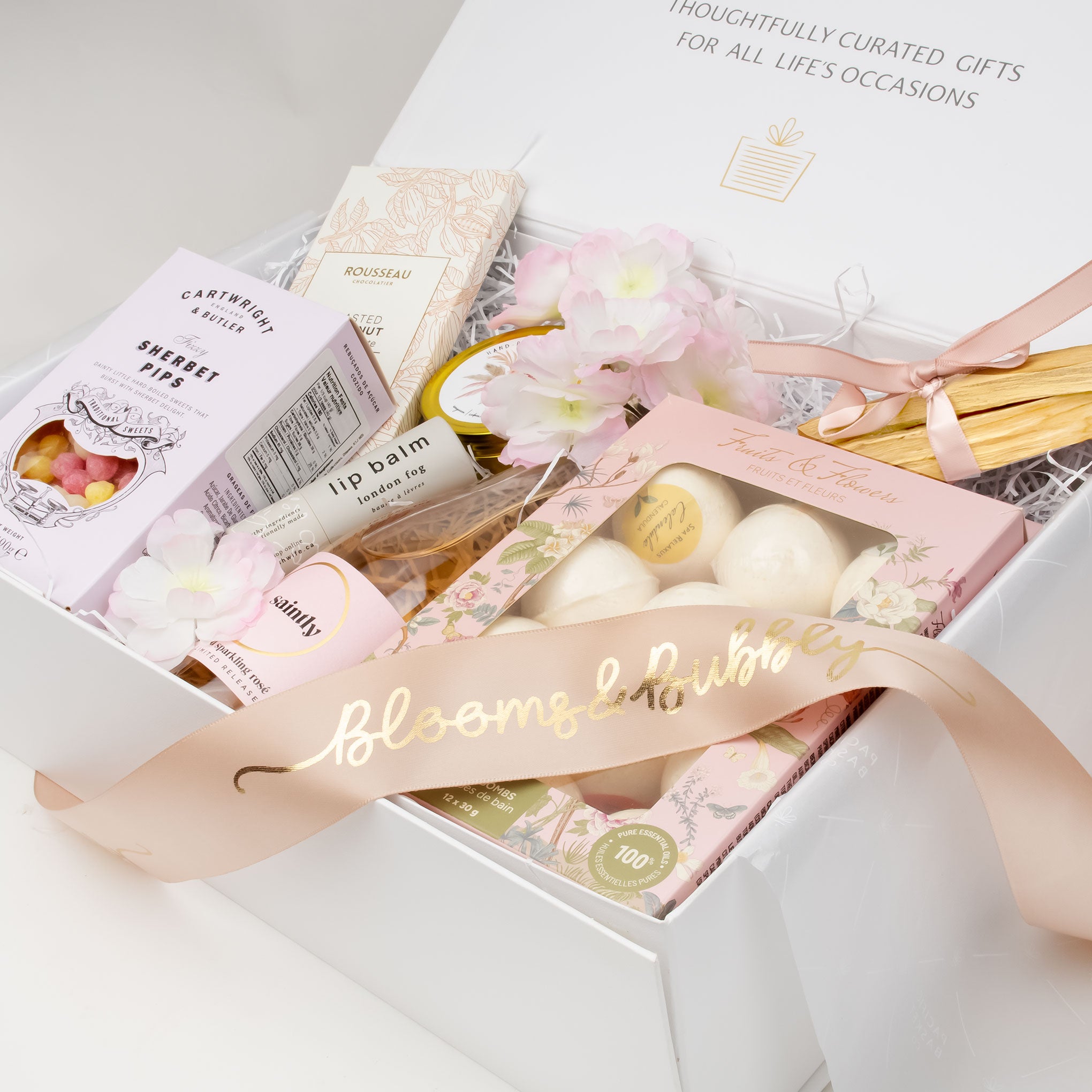 Blooms and Bubbly' spring gift basket featuring a bottle of bubbly, luxury bath bombs, a Palo Santo bundle, artisanal chocolate, lip balm and a premium soy candle, elegantly arranged with a satin bow.